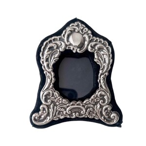 Könnte beinhalten: Ein silberfarbener Bilderrahmen mit einem verzierten, geprägten Metallrand, der komplizierte Schnörkel, florale Motive und ein glattes zentrales Wappen aufweist. Der Rahmen umschließt ein dunkles, ovales Zentrum und ist auf schwarzem Samt montiert.