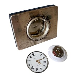 May include: A vintage silver-toned clock with a square base and a circular opening. The clock face is white with black Roman numerals and gold hands. The text "KITNEY & CO LONDON" is visible. A white plastic component and a battery are also present.
