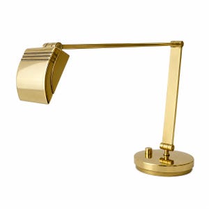 May include: A gold-colored desk lamp with a rounded base, adjustable arm, and a rectangular shade. The shade has horizontal lines. The lamp has a classic, Art Deco design.