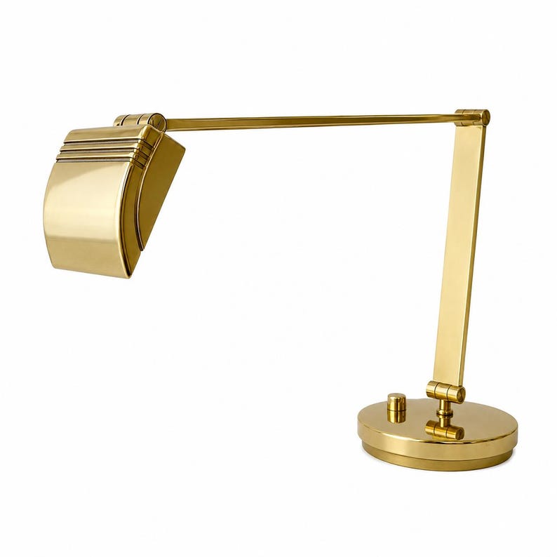 May include: A gold-colored desk lamp with a rounded base, adjustable arm, and a rectangular shade. The shade has horizontal lines. The lamp has a classic, Art Deco design.