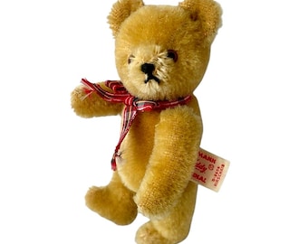 May include: A golden-brown mohair teddy bear with dark eyes and a stitched nose. The bear wears a red and black plaid ribbon around its neck. A white fabric tag is attached to the side, featuring red text that reads Hermann Teddy Original.