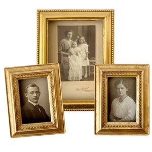 Pode incluir: Três molduras de fotos douradas com detalhes ornamentados. As molduras estão dispostas em forma de triângulo. A moldura superior contém uma fotografia em preto e branco de uma mulher e duas crianças. A moldura da esquerda contém uma fotografia em preto e branco de um homem. A moldura da direita contém uma fotografia em preto e branco de uma mulher.