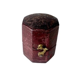 May include: A small, hexagonal jewelry box with a textured, deep burgundy exterior finish. The box features a dark, mottled lid and a central brass-colored metal latch mechanism on the front that secures the two halves of the container together.