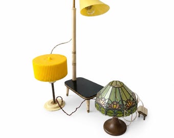 Op de afbeelding: Een verzameling vintage lampen, waaronder een gele, kegelvormige vloerlamp, een kleine gele tafellamp en een tafellamp met gebrandschilderd glas met een groen en bloemenpatroon. Op een witte achtergrond.