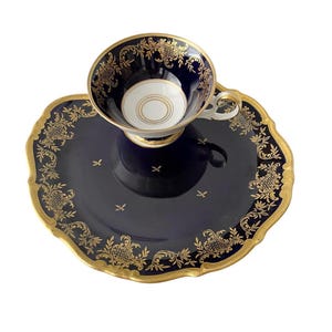 May include: A dark blue porcelain teacup and matching saucer set featuring intricate gold filigree patterns along the scalloped edges. The interior of the cup is white with gold circular accents, and the handle is white with gold trim detailing.