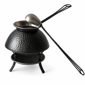 Puede incluir: Un quemador de incienso de metal negro con textura martillada, borde plateado y un mango largo y delgado. El quemador se asienta sobre una base de tres patas. La tapa está abierta, revelando el interior. El mango mide unos 46 cm.