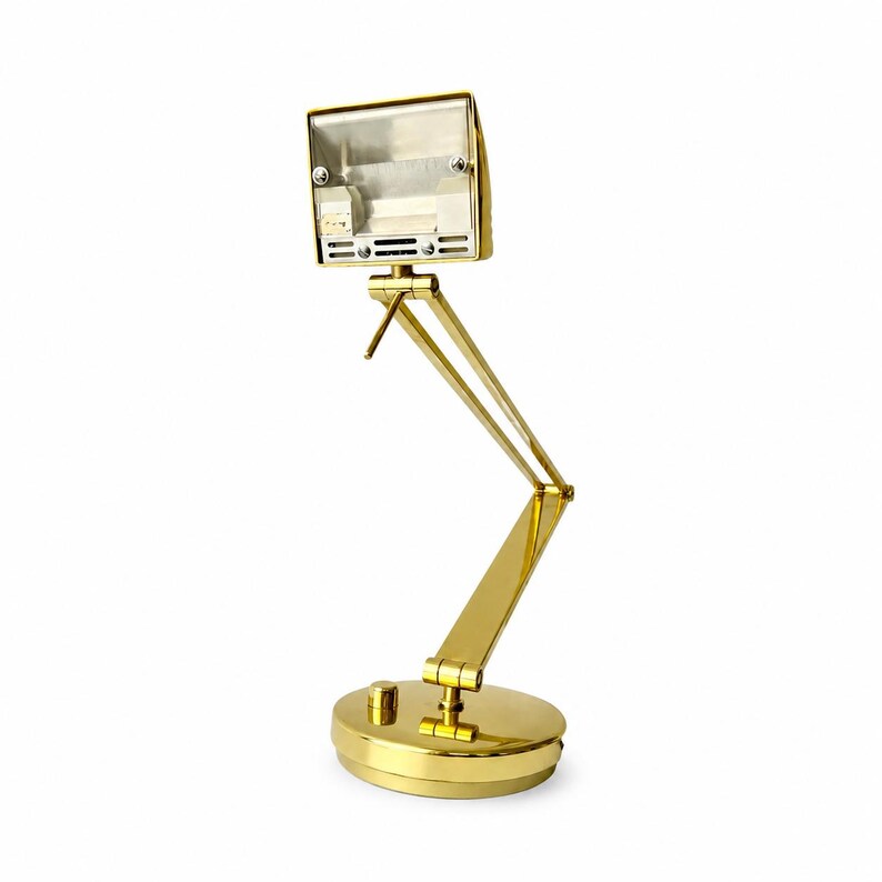 May include: A gold-colored desk lamp with a rectangular head and an adjustable arm. The lamp has a round base with a small button and a pivoting head. The lamp is made of metal and has a shiny finish.