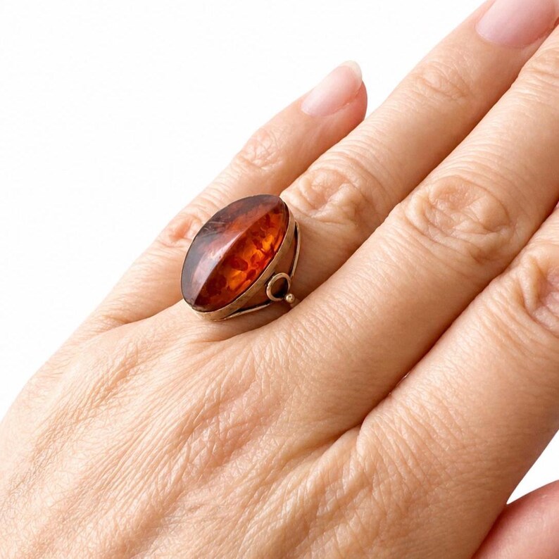 May include: An oval-shaped amber ring with a gold-colored setting. The amber stone is a rich, translucent brown with a polished surface. The ring is worn on a finger, showcasing its size and design. A classic piece of jewelry.