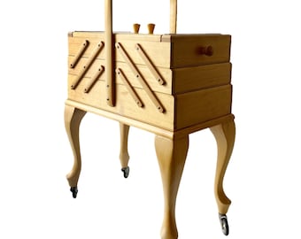 May include: A wooden sewing box with a hinged lid and a drawer. The box has a handle and sits on four legs with wheels. The box is made of light wood and has a natural finish.