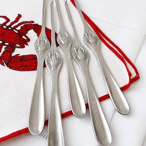 May include: Five stainless steel seafood forks with lobster head designs, resting on a white linen napkin with a red embroidered lobster and trim. The forks have a long, slender design with two tines.