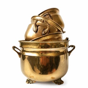Puede incluir: Una pila de ollas y sartenes antiguas de latón. La olla más grande tiene asas ornamentadas y descansa sobre tres pies decorativos. Las ollas más pequeñas tienen asas curvas. El latón tiene un tono dorado cálido, que refleja la luz.