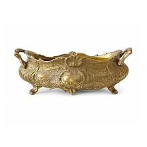 Puede incluir: Un cuenco alargado de metal dorado con patrones en relieve, un borde festoneado y dos asas laterales. El recipiente se apoya sobre cuatro pies pequeños y curvados, mostrando una superficie texturizada con volutas y motivos de conchas.