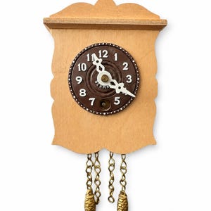 May include: A small, wooden cuckoo clock with a light brown finish. The clock face is dark brown with white numbers and hands. Two gold-colored pinecone weights hang from chains below.