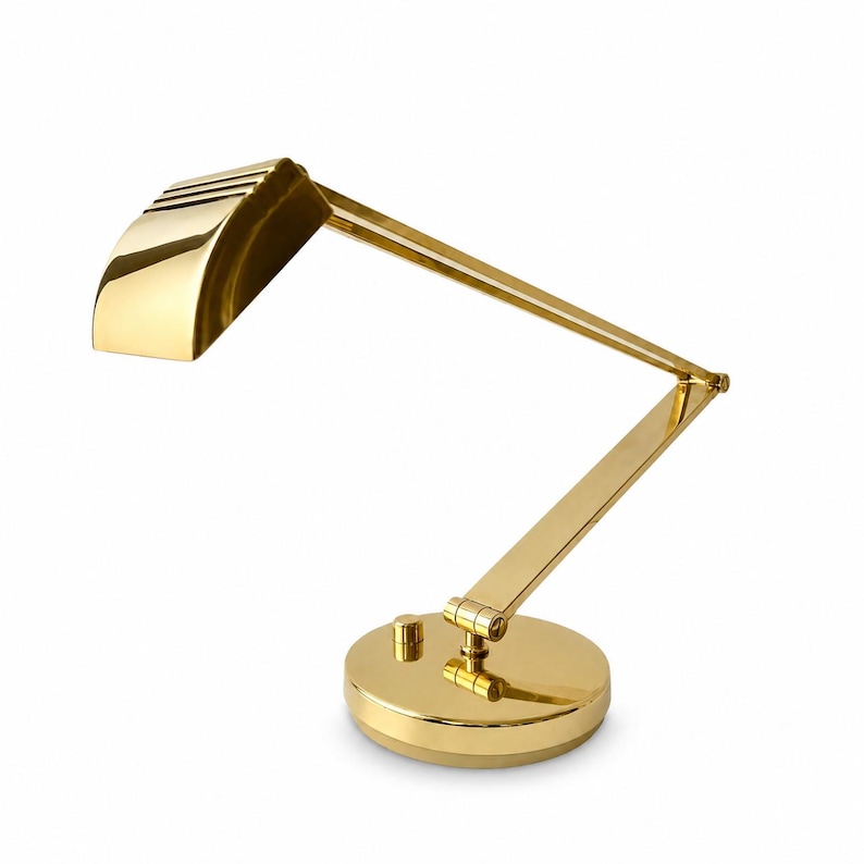 May include: A gold-colored desk lamp with an adjustable arm and a round base. The lamp has a rectangular shade with horizontal slits and a small on/off switch. The lamp is made of metal and has a shiny finish.