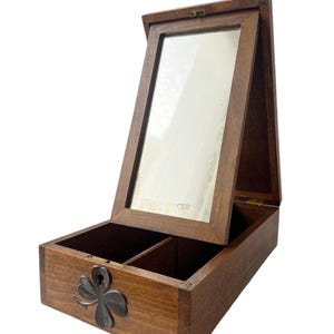 May include: A wooden jewelry box with a hinged, framed mirror. The box is stained a warm brown and features a decorative metal shamrock on the front. The interior is divided into two compartments.