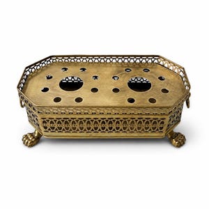 May include: A gold-toned, octagonal serving tray with a decorative pierced design. The tray has two large circular openings and smaller holes on the top surface. It features ornate detailing, including a patterned border and claw feet.