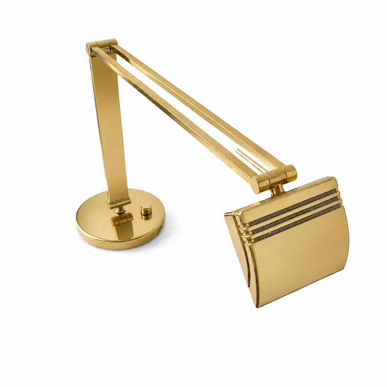 May include: A gold-colored desk lamp with an adjustable arm and a round base. The lamp has a rectangular shade with three horizontal lines and a small on/off switch. The lamp is made of metal and has a shiny finish.