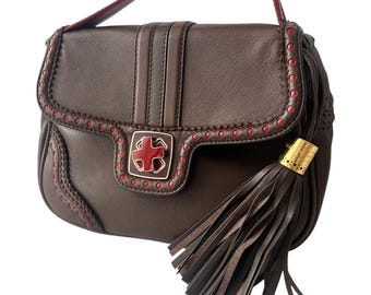 May include: A brown leather handbag with a red handle and trim. The bag features a decorative red and gold emblem on the front and a long tassel. The bag has a structured shape and a top handle.