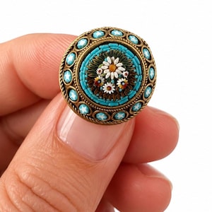 Può includere: Una spilla rotonda caratterizzata da un design centrale a mosaico con margherite bianche dal centro giallo su sfondo scuro, circondata da un bordo di piccole tessere turchesi e una cornice esterna in ottone con accenti azzurri a forma di stella.