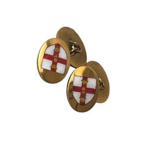 Puede incluir: Un par de gemelos dorados de forma ovalada. Cada gemelo presenta un escudo blanco con una cruz roja y detalles dorados. Probablemente vintage o antiguo.