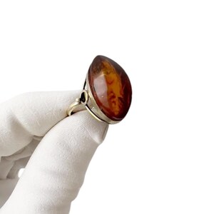 May include: A silver ring featuring a large, teardrop-shaped amber stone. The amber has a warm, reddish-brown hue with natural inclusions. The ring's setting is a simple, elegant design.