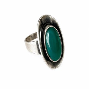 Puede incluir: Un anillo de plata con una piedra verde grande, lisa y de forma ovalada en el centro. La banda de metal tiene un acabado oxidado y desgastado, y la piedra está rodeada por un marco de plata ancho y cóncavo que crea una silueta ovalada alargada.