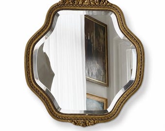 May include: An antique-style mirror with a gold-coloured, decorative frame. The mirror has a distinctive, scalloped edge and ornate detailing. The reflection shows a room with paintings and a window, creating a classic, elegant look.