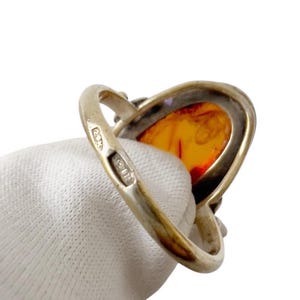 May include: Silver ring featuring a large, oval amber stone with a warm, orange hue. The ring's band is a polished silver, and the amber stone is set in a silver bezel. The ring is held by a gloved hand.