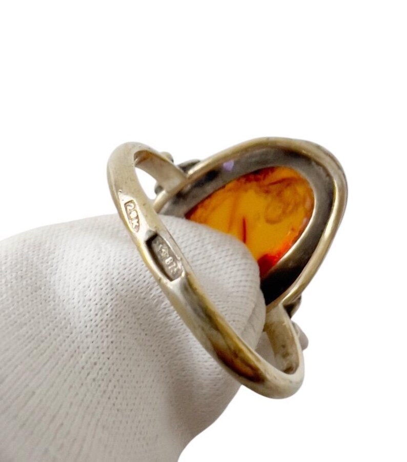 May include: Silver ring featuring a large, oval amber stone with a warm, orange hue. The ring's band is a polished silver, and the amber stone is set in a silver bezel. The ring is held by a gloved hand.