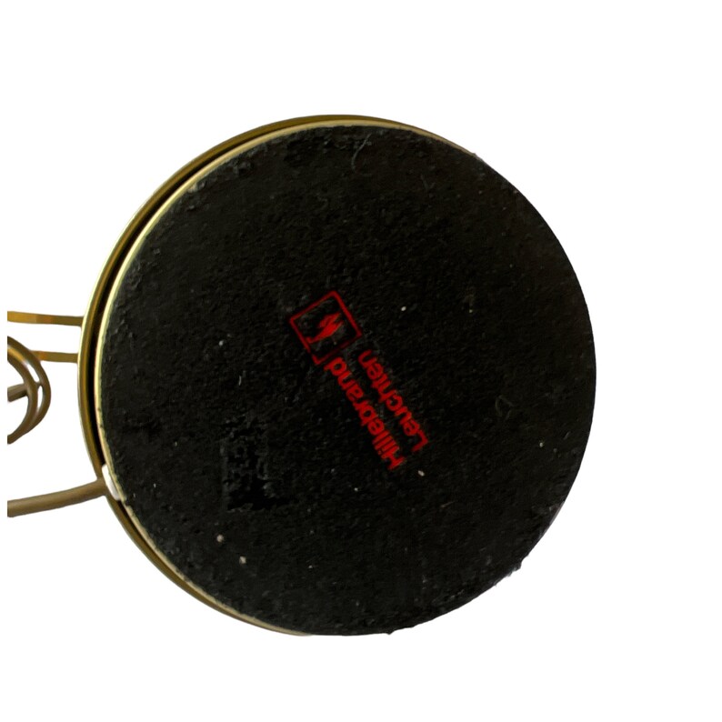 May include: Close-up of the bottom of a gold-colored lamp. The base is a black, circular shape with the red text "Hillebrand Leuchten" printed on it. The lamp's metal frame is visible.