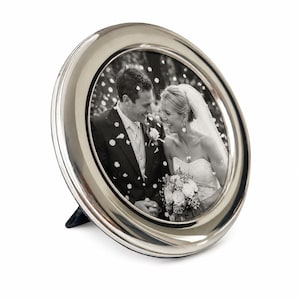 May include: A round, polished silver-tone metal picture frame with a smooth, reflective border. The frame contains a black and white photograph of a couple in wedding attire, with the person on the left in a suit and the person on the right in a gown.