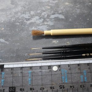 Può includere: Quattro pennelli, tre con manici neri e uno con manico crema, sono disposti su una superficie grigia. Un righello con segni in centimetri è visibile in primo piano.