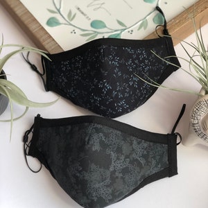 May include: Two black face masks with different floral patterns. The top mask has a light blue floral pattern and the bottom mask has a dark gray floral pattern.