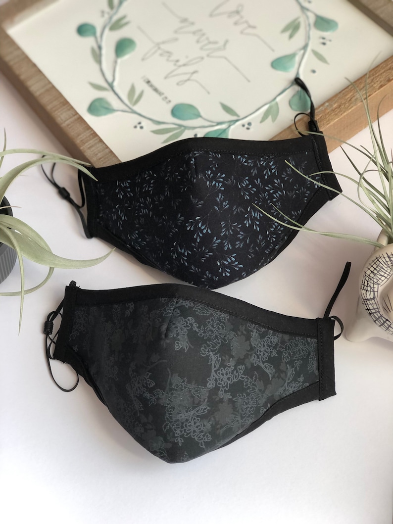 May include: Two black face masks with different floral patterns. The top mask has a light blue floral pattern and the bottom mask has a dark gray floral pattern.