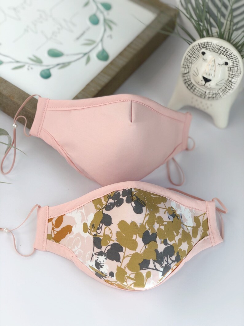 Op de afbeelding: Twee roze gezichtsmaskers met verstelbare oorlussen. E&eacute;n masker is effen roze, de andere heeft een bloemenpatroon in goud, grijs en wit op een roze achtergrond. De maskers zijn ontworpen voor comfort en bescherming.