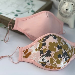 Op de afbeelding: Twee gezichtsmaskers. E&eacute;n is effen lichtroze, de andere heeft een bloemenpatroon in roze, groene en grijze tinten. Beide hebben verstelbare oorlussen. De maskers liggen op een wit oppervlak, met een ingelijste foto en een decoratieve leeuw op de achtergrond.