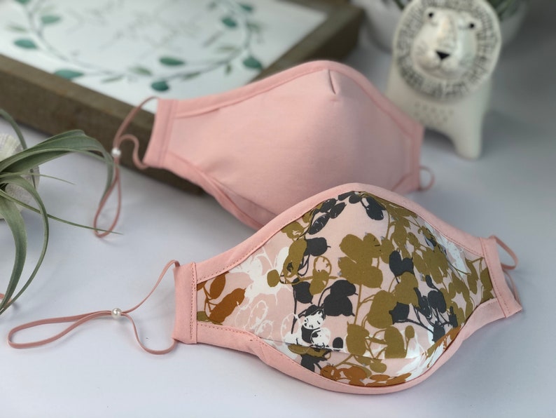 Op de afbeelding: Twee gezichtsmaskers. E&eacute;n is effen lichtroze, de andere heeft een bloemenpatroon in roze, groene en grijze tinten. Beide hebben verstelbare oorlussen. De maskers liggen op een wit oppervlak, met een ingelijste foto en een decoratieve leeuw op de achtergrond.