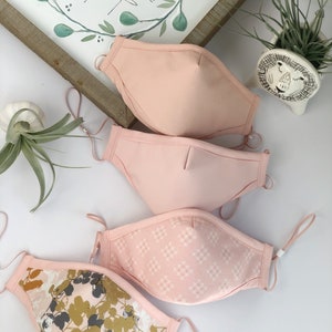 Op de afbeelding: Vier roze gezichtsmaskers. Twee zijn effen, &eacute;&eacute;n heeft een wit geometrisch patroon en de laatste heeft een bloemenontwerp met gouden en groene accenten. Elk masker heeft verstelbare oorlussen.