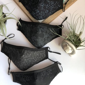 May include: Four black face masks with different patterns. The masks are made of fabric and have adjustable ear loops. The patterns include a floral design, a geometric design, a lace design, and a small leaf design.