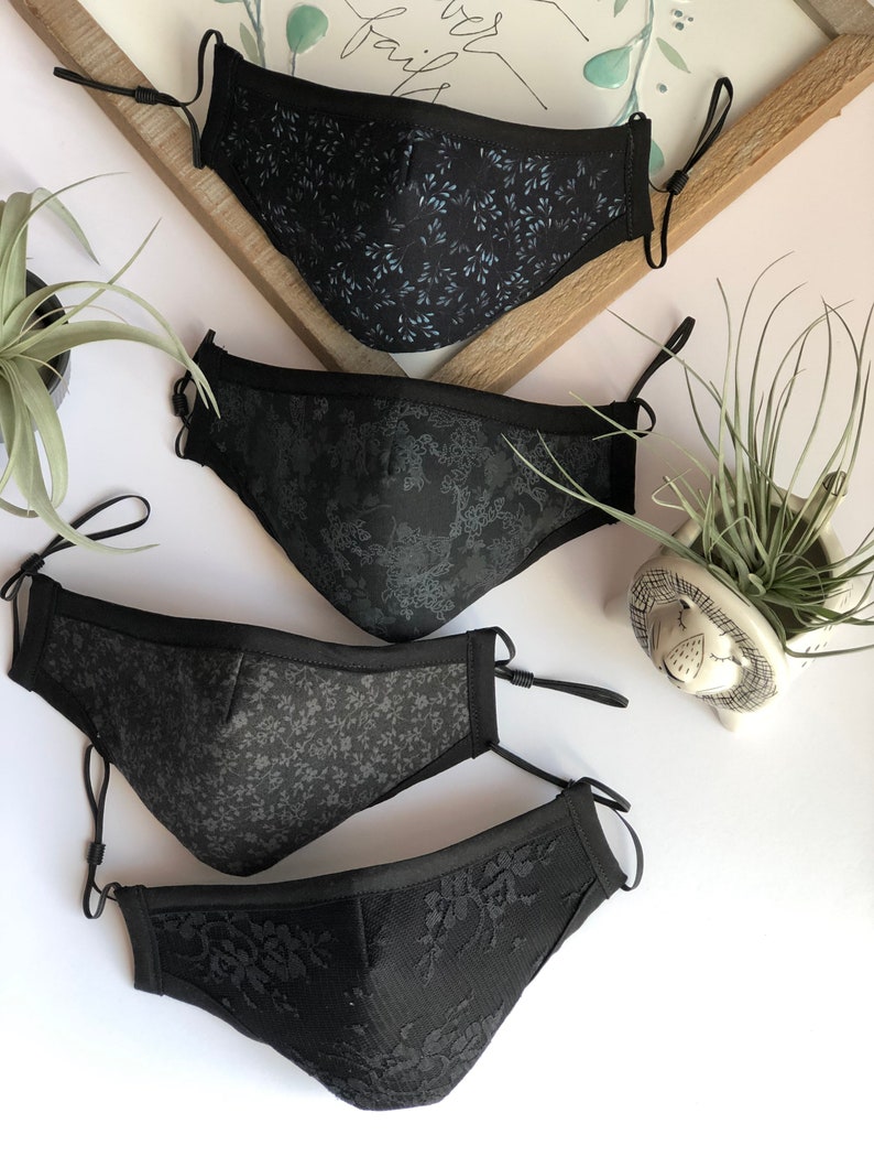 May include: Four black face masks with different patterns. The masks are made of fabric and have adjustable ear loops. The patterns include a floral design, a geometric design, a lace design, and a small leaf design.