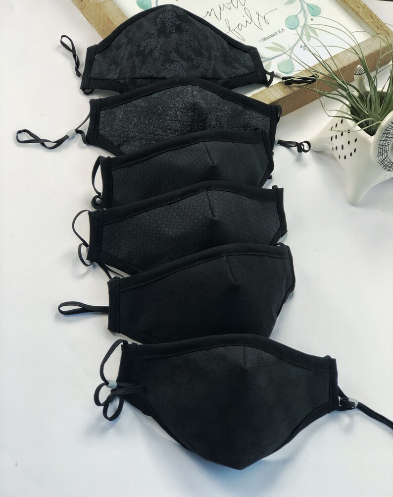 May include: A stack of five black face masks with different patterns. The masks are made of a soft, breathable fabric and have adjustable ear loops.