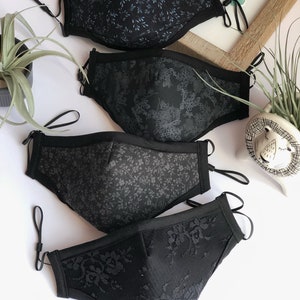 May include: Four black face masks with different patterns. The top mask has a small blue floral pattern, the second mask has a larger floral pattern, the third mask has a small floral pattern, and the bottom mask has a lace pattern. The masks are all made of fabric and have elastic ear loops.