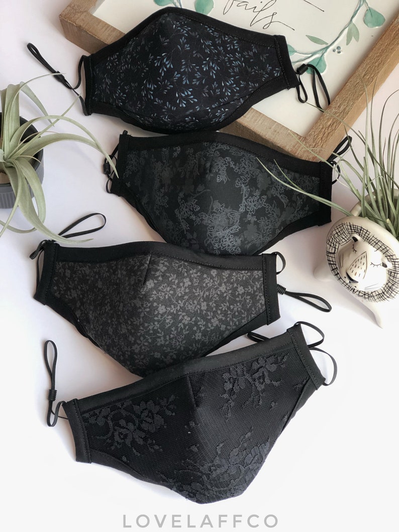 May include: Four black face masks with different patterns. The top mask has a small blue floral pattern, the second mask has a larger floral pattern, the third mask has a small floral pattern, and the bottom mask has a lace pattern. The masks are all made of fabric and have elastic ear loops.