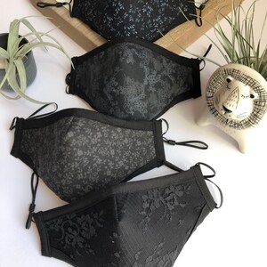 May include: Four black face masks with different patterns. The masks are made of fabric and have elastic ear loops. The patterns include a floral design, a small leaf design, a larger leaf design, and a lace design.