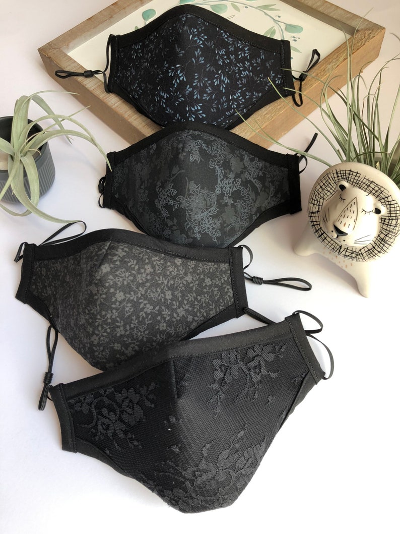 May include: Four black face masks with different patterns. The masks are made of fabric and have elastic ear loops. The patterns include a floral design, a small leaf design, a larger leaf design, and a lace design.