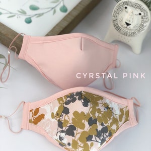 Op de afbeelding: Twee gezichtsmaskers. Het bovenste masker is effen lichtroze, met het label "CYRSTAL PINK". Het onderste masker heeft een roze bloemenpatroon met gouden en grijze bladeren, met het label "PINK SAKURA". Beide maskers hebben verstelbare oorlussen.