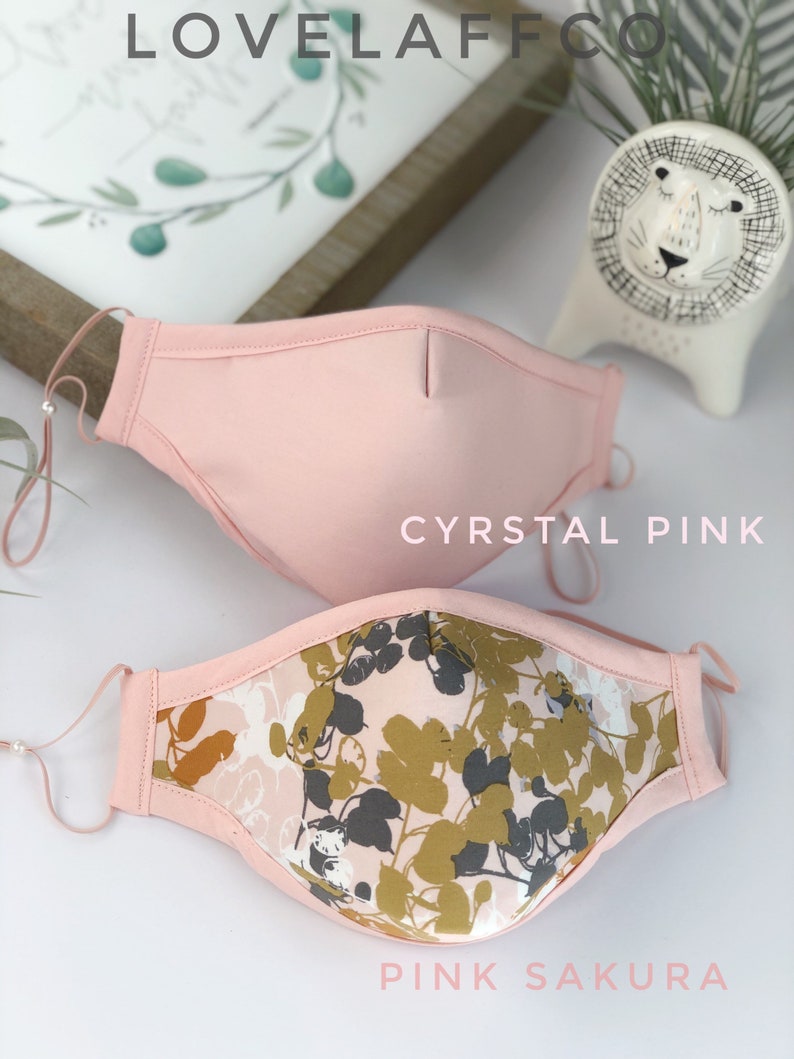 Op de afbeelding: Twee gezichtsmaskers. Het bovenste masker is effen lichtroze, met het label "CYRSTAL PINK". Het onderste masker heeft een roze bloemenpatroon met gouden en grijze bladeren, met het label "PINK SAKURA". Beide maskers hebben verstelbare oorlussen.
