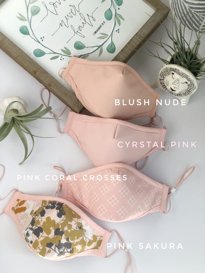 Op de afbeelding: Vier roze gezichtsmaskers met verstelbare oorlussen. De maskers zijn gelabeld met "Blush Nude", "Cyrstal Pink", "Pink Coral Crosses" en "Pink Sakura". Op de achtergrond een ingelijste print met de woorden "love never fails".