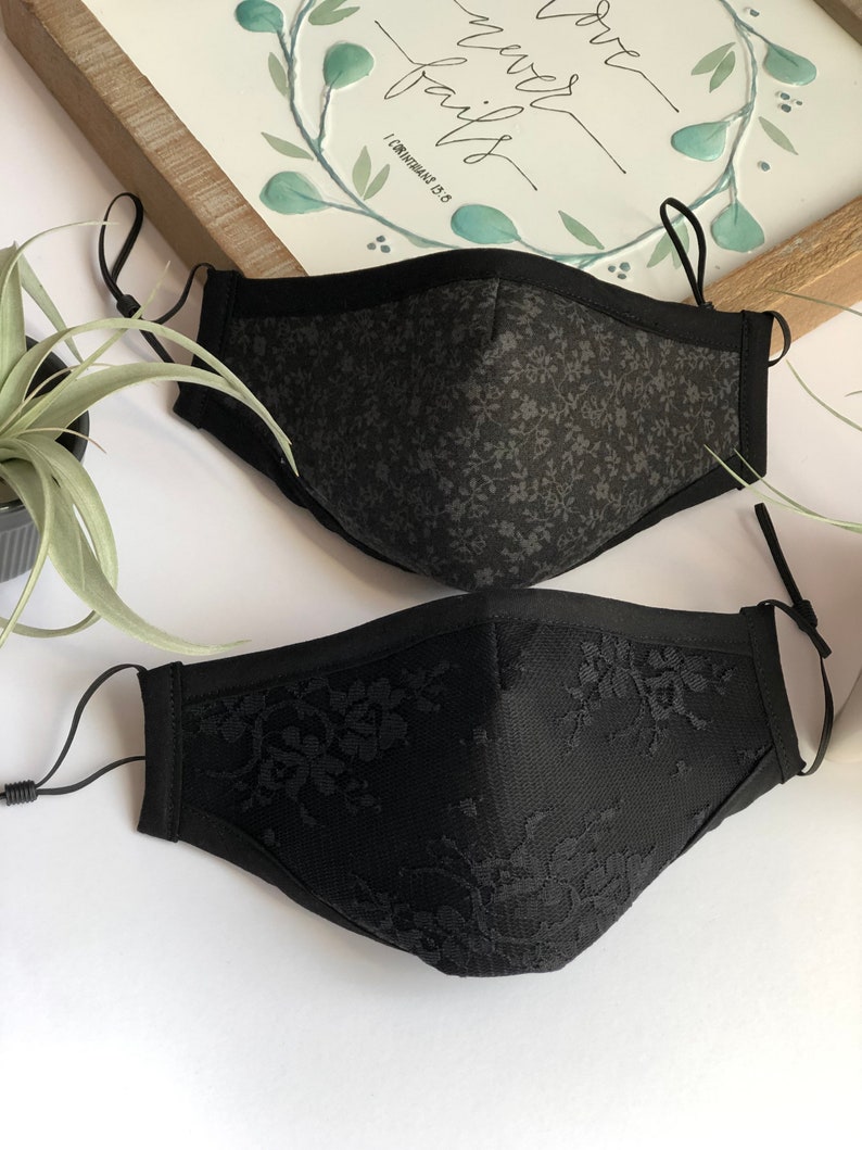 May include: Two black face masks. One mask is made of a black fabric with a small floral pattern. The other mask is made of black lace.