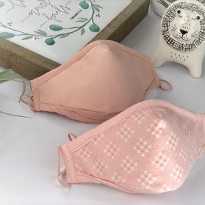 Op de afbeelding: Twee roze gezichtsmaskers met verstelbare oorlussen. E&eacute;n masker is effen roze en het andere heeft een wit en roze kruispatroon. De maskers zijn gemaakt van stof en zijn ontworpen om de neus en mond te bedekken.